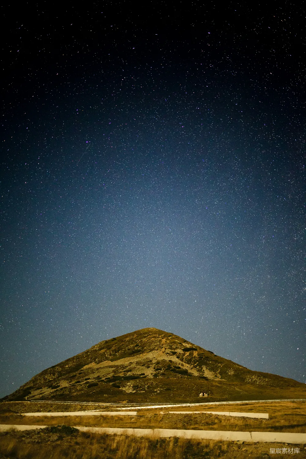 这张图片展示了夜晚的夜空，星星点点，明亮而璀璨。前景是一座起伏的山丘，山丘的轮廓在星光的映衬下显得格外清晰。地面上有白色的条形结构，可能是道路或建筑的一部分。整体画面宁静而美丽，适合用作自然风光的背景素材。
