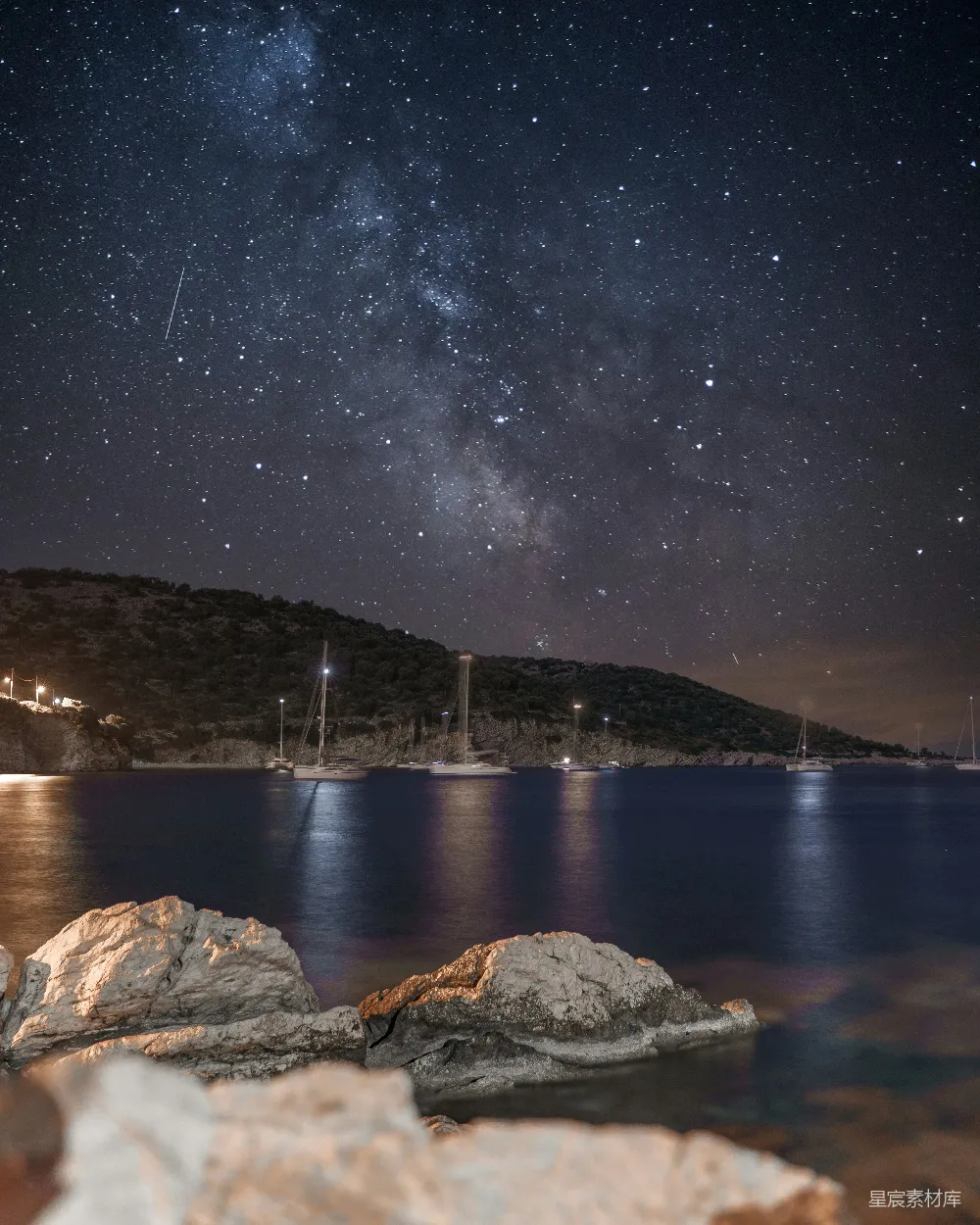 这张图片展示了夜幕下的海岸景象。满天的星星在深蓝色的夜空中闪烁，银河清晰可见。海面上停泊着几艘船只，岩石在前景中显得格外突出。整体画面宁静而美丽，适合用于展示自然风光或旅行摄影。