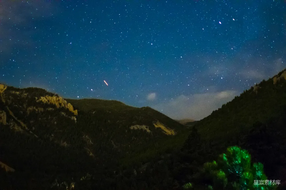 这张图片展示了宁静的夜空下的山谷景色。深蓝色的夜空布满了星星，山谷中覆盖着茂密的绿色植被，远处的山峦在夜色中若隐若现。地平线处有一道流星划过，增添了画面的动感与神秘感。整体氛围宁静而深邃，适合用作自然风光或宁静夜晚的背景图。
