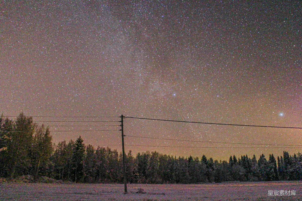 这张图片展示了一个宁静的冬季夜晚，星空布满了无数的星星，天空呈现出柔和的紫色和黄色渐变。地面上覆盖着一层薄雪，远处是一片茂密的森林，前景中有一根电线杆和电线，增添了画面的层次感。整体氛围静谧而美丽，非常适合用于展示自然风光和宁静的冬夜场景。