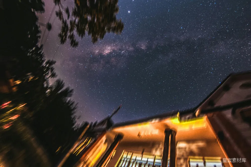 这张图片展示了夜晚的景象，星空璀璨，银河清晰可见。画面中有一些建筑物，灯光温暖，与天空的深邃形成对比。树木的剪影和建筑的轮廓增添了画面的层次感。适合用作夜景摄影、自然风光或城市夜景的素材。