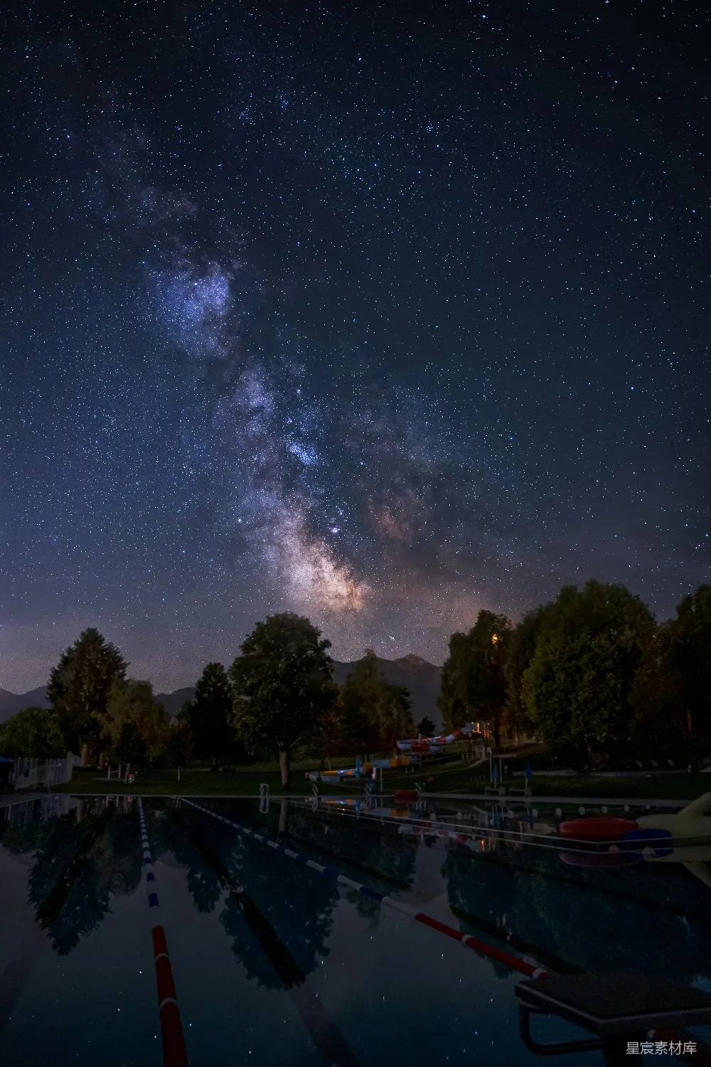 这张图片展示了一个宁静的夜晚，星空璀璨，银河清晰可见，倒映在泳池中。泳池周围是茂密的树木和远处的山脉，营造出一种宁静而神秘的氛围。适合用于展示自然美景和宁静的夜晚场景。