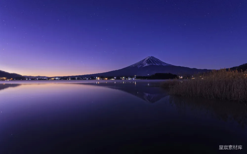 这张图片展示了一个宁静的夜晚，富士山巍峨挺立，倒映在平静的湖水中，天空中布满了星星，远处的城市灯光点缀其中。整体画面宁静而美丽，适合用作自然风景或旅行主题的背景素材。