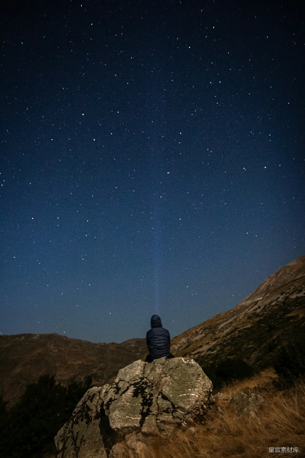 图片中，一位旅人坐在岩石上，仰望满天星辰。天空深邃而宁静，星星点缀其间，与下方的山脉形成对比。画面传达出一种宁静与探索的精神，适合用作旅行、自然、宁静等主题的背景或标题。