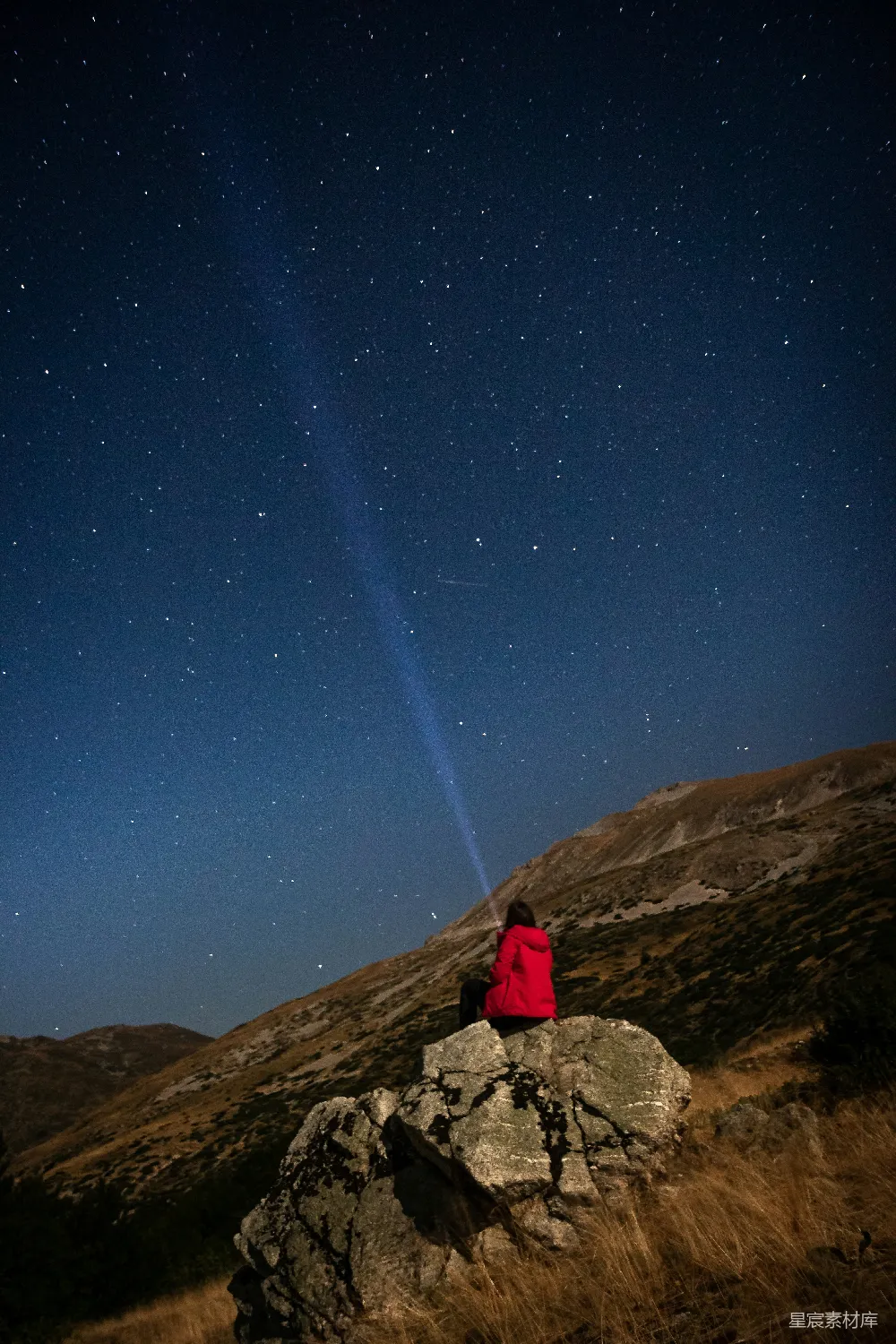 一位身穿红色外套的旅人在山顶岩石上仰望星空，手中似乎握着或指向一条明亮的光束。背景是深蓝色的夜空，点缀着无数星星，远处的山丘轮廓在夜色中若隐若现。这张照片捕捉了宁静与神秘并存的时刻，适合用作旅行、自然、摄影等主题的背景素材。