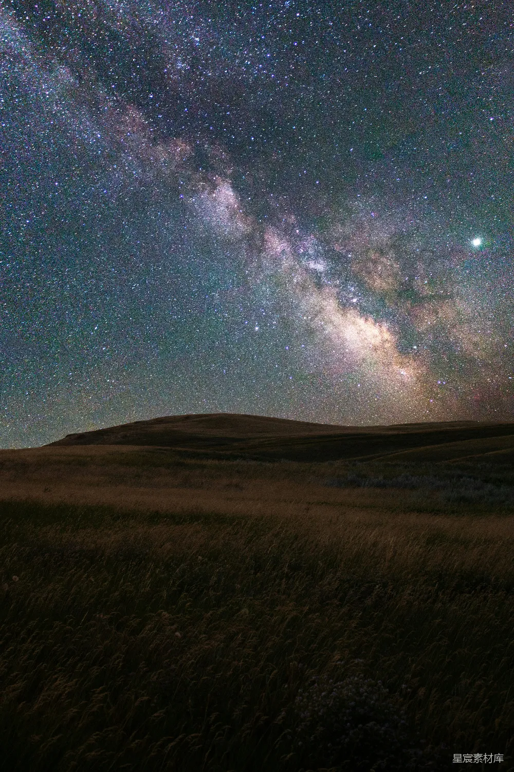 这张图片展示了夜晚的天空，银河横跨天际，星星点点，背景是宁静的田野。画面中，地平线上的山脉轮廓清晰，而近处的草地在微弱的光线下若隐若现，营造出一种宁静而神秘的氛围。适合用于自然风光、星空摄影和宁静主题的背景素材。