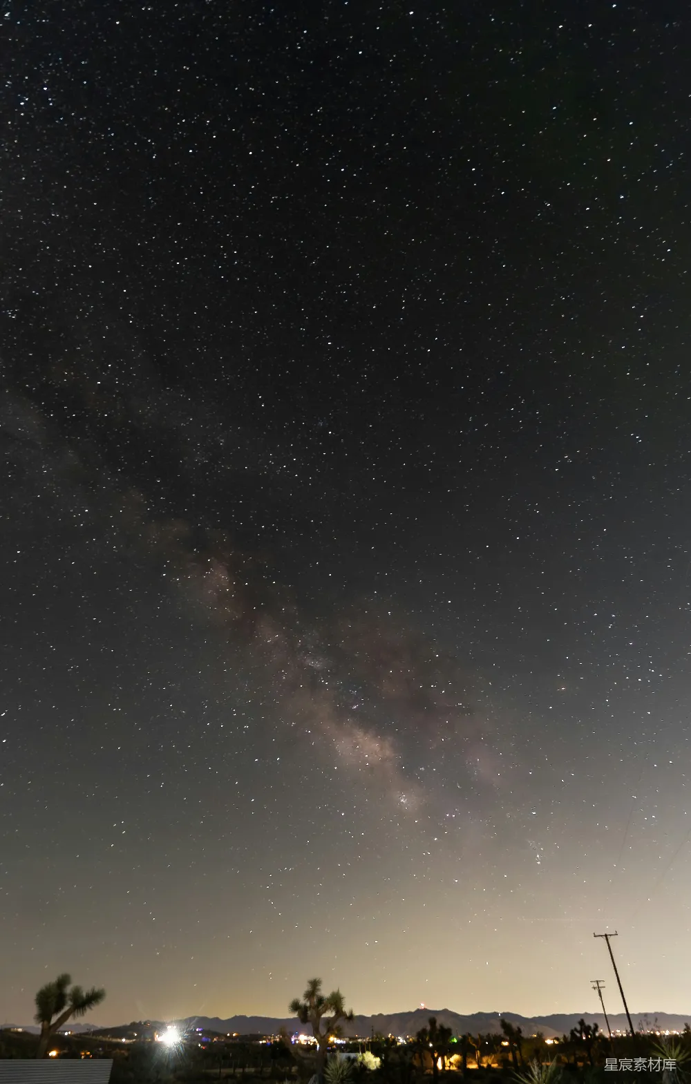 这张图片展示了夜空中壮丽的银河景象，银河在星空下清晰可见，背景是宁静的沙漠夜景，点缀着稀疏的树木和远处的灯光，营造出一种神秘而宁静的氛围。适合用于自然风光、天文摄影、宁静时刻的背景或壁纸。