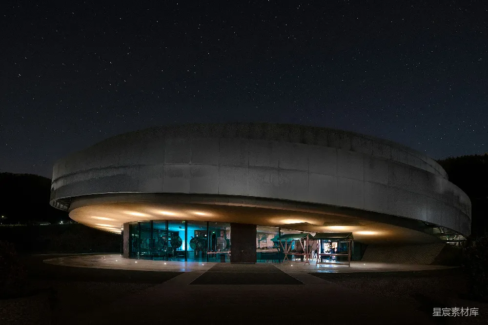 这张图片展示了一座独特的现代建筑在夜晚的景象。建筑外形圆润，顶部呈浅色，底部则被灯光照亮，显得格外引人注目。背景是深邃的夜空，点缀着无数星星，营造出一种宁静而神秘的氛围。适合用作城市夜景、现代建筑或夜间摄影的素材。