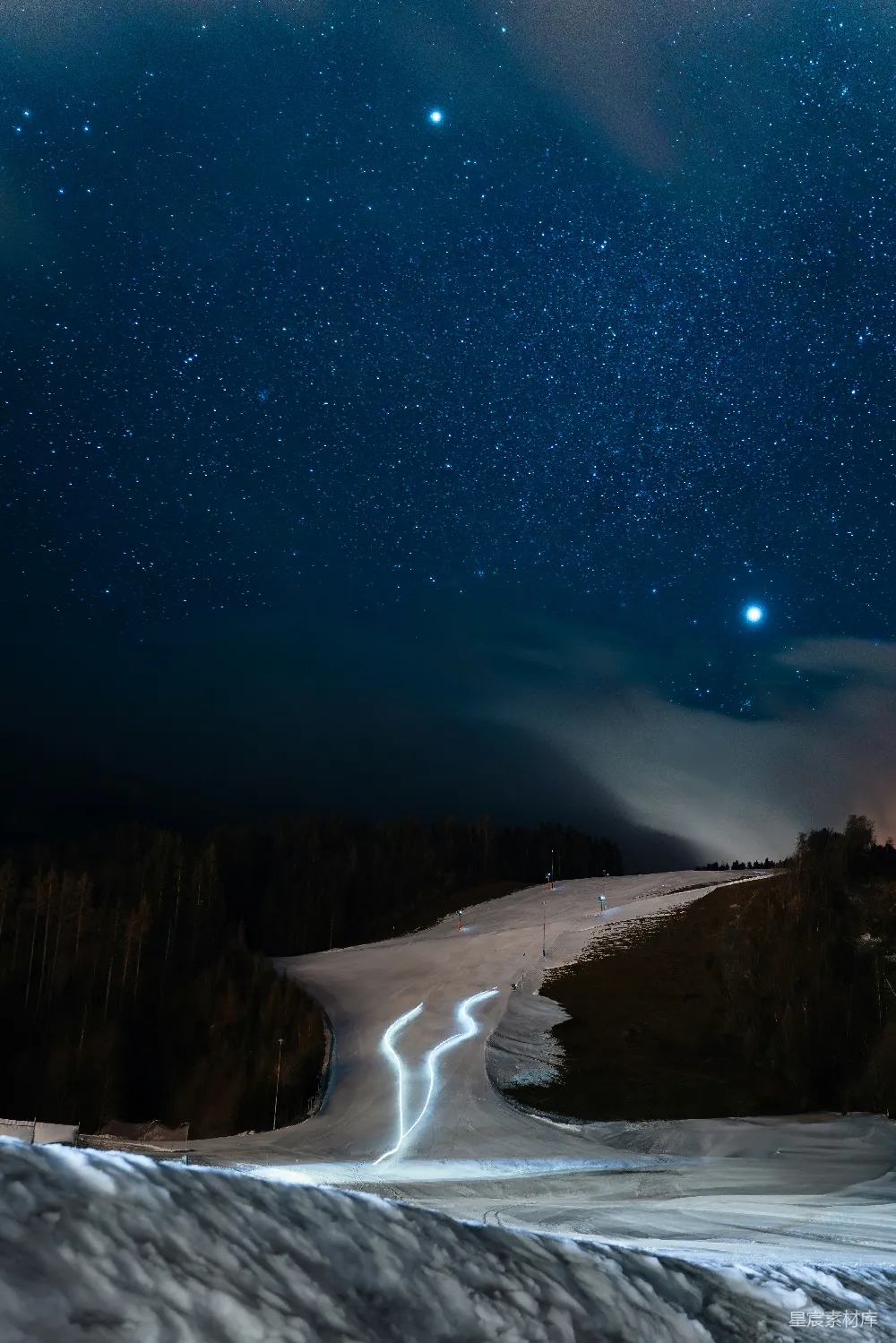 这张照片展示了夜晚宁静的滑雪道，星空璀璨，几颗明亮的星星点缀在夜空中。滑雪道上，两条曲线清晰可见，仿佛时间在此刻静止。周围是深邃的森林，远处的灯光勾勒出道路的轮廓，整体氛围宁静而神秘。适合用作自然美景、夜景摄影、冬季运动的背景素材。
