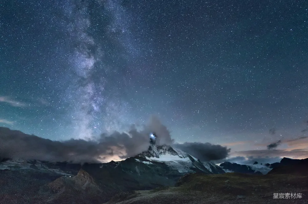 这张图片展示了夜空中繁星点点，银河横跨天际，背景是雪山覆盖的山峰，山峰上似乎有一处光源，与周围的黑暗形成鲜明对比，营造出一种神秘而宁静的氛围。适合用于展示自然风光、摄影艺术以及旅行探险的主题。