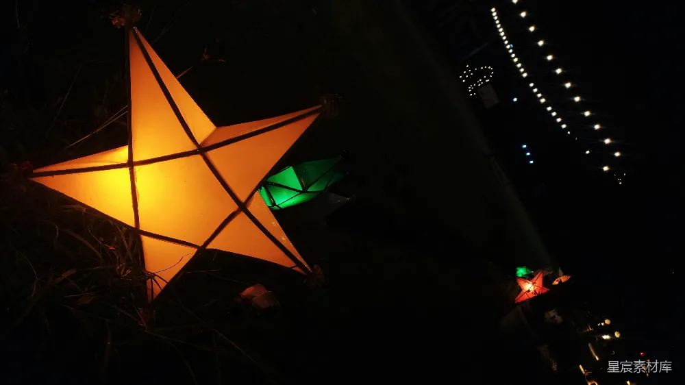 图片展示了一组夜晚悬挂的星星灯饰，橙色和绿色的灯光在黑暗中显得格外耀眼，营造出一种节日和温馨的氛围。背景中还隐约可见一排小灯串，增添了画面的层次感和节日气氛。适合用于装饰户外活动、节日庆典或夜晚的休闲场景。