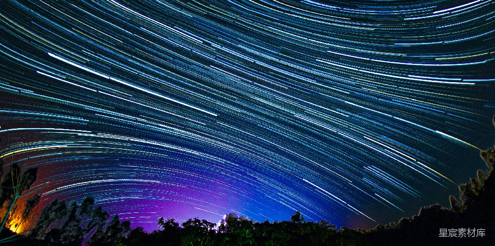 这张图片展示了在夜晚，星轨在夜空中形成的一道道光带。色彩以蓝色和紫色为主，背景是深邃的夜空，前景有树木的剪影。这是一种长时间曝光拍摄的星空照片，展示了地球自转过程中星星的轨迹。这种场景适合用作壁纸、背景图，或者用于表现宁静、神秘或探索主题的