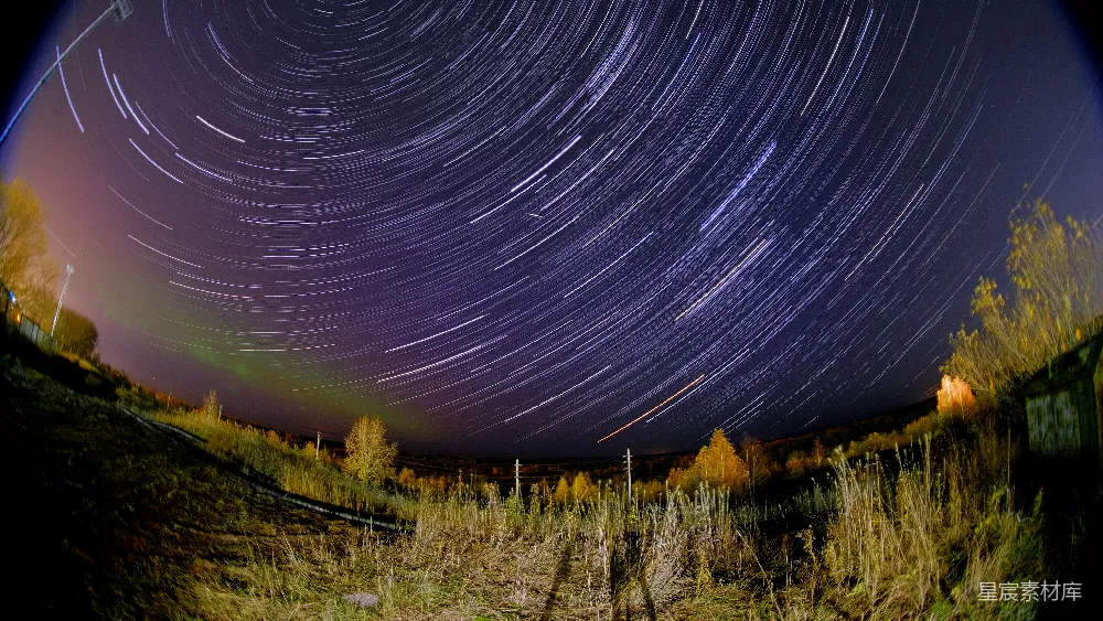 这张图片展示了夜空中星轨的美丽景象。星轨是由长时间曝光拍摄得到的，显示了星星在夜空中移动的轨迹。图片中可以看到远处的树木和一些灯光，增添了画面的深度和细节。整体效果非常壮观，适合用于自然风光、星空摄影或夜景主题的图片。