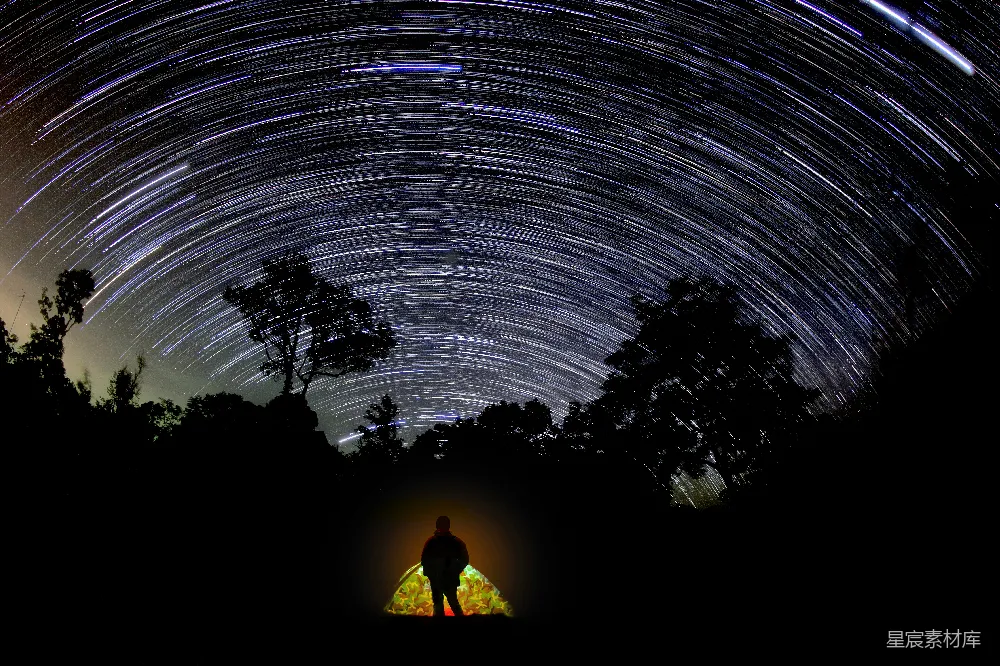 这张图片展示了一片星空下的露营场景。夜空中，星星的轨迹在夜幕上形成了美丽的星轨，而帐篷内的灯光则照亮了露营者，营造出一种宁静而神秘的氛围。适合用于展现自然美景、露营体验或夜景摄影的主题。