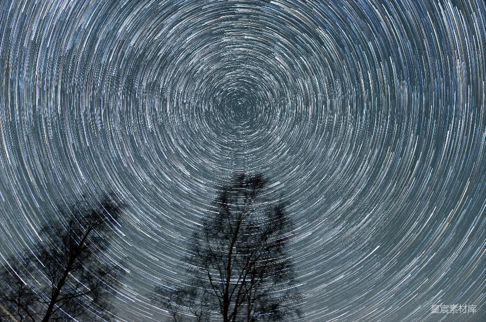 这张图片展示了在长曝光拍摄下，夜空中星星轨迹形成的旋转图案。中心的旋转轨迹由恒星的缓慢移动和相机的固定拍摄位置共同作用产生，形成了一个美丽的圆形图案。底部的树木剪影为画面增添了深度，背景的星空则显得更加神秘。这张照片适合用于天文、自然风光和