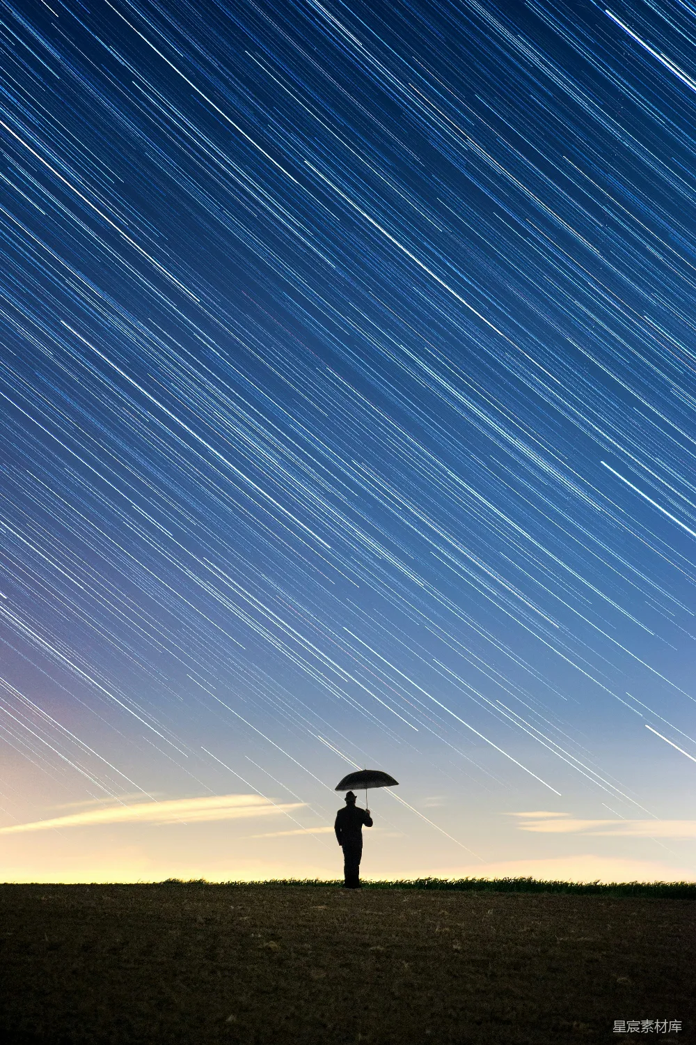 图片中，一个人在星空下撑着伞，背景是壮观的星轨，营造出宁静而神秘的氛围。星轨的线条与地平线的结合，展现了时间与空间的流动感，适合用作宁静、自然、星空等主题的背景素材。
