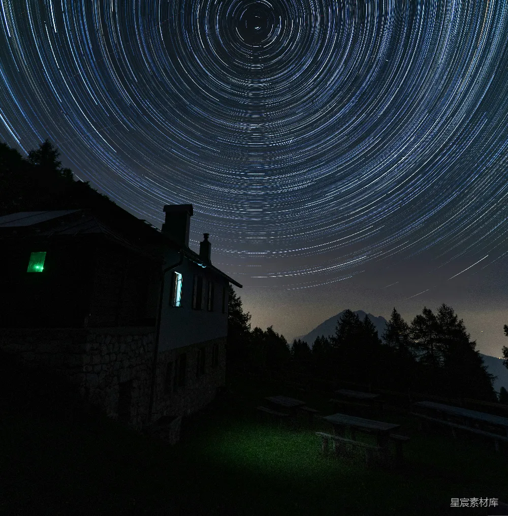 这张图片展示了一个夜晚的场景，天空中布满了美丽的星轨。背景是一座石制的小屋，周围是茂密的树木和远处的山脉。星轨的轨迹形成了一个完美的圆形，营造出一种宁静而神秘的氛围。图片整体色调偏暗，但星轨的光带使得画面非常引人注目。适合用于自然风光、旅行