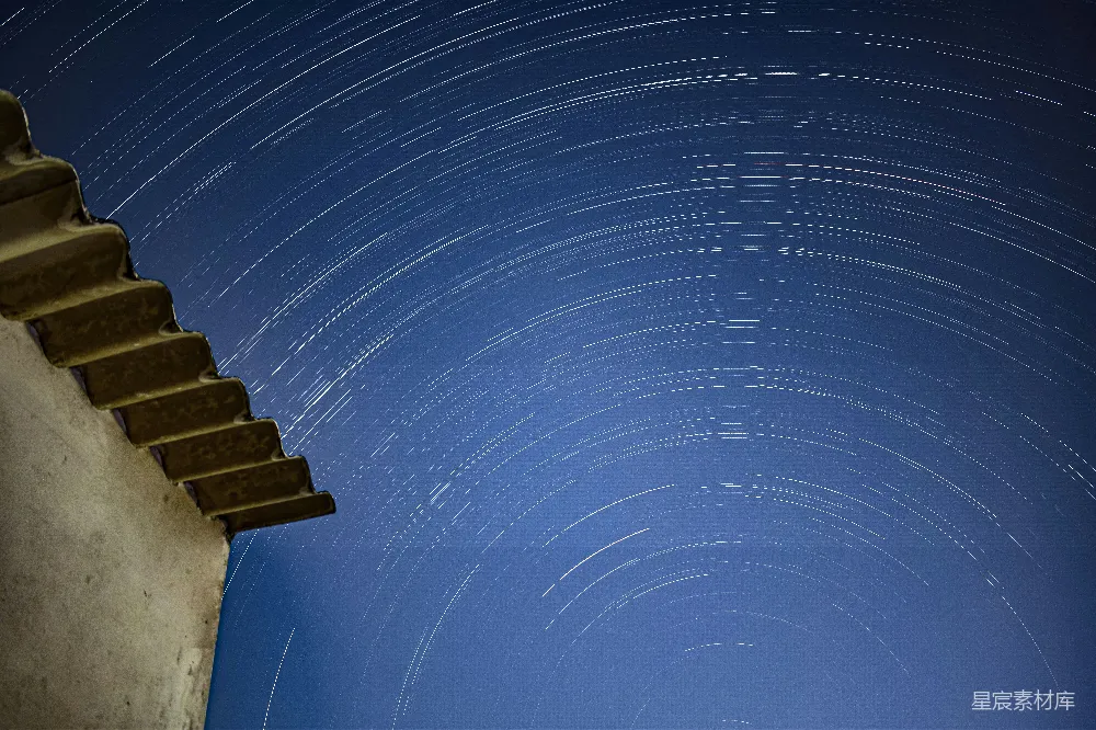 这张图片展示了夜晚的星空，星轨在夜空中形成美丽的螺旋状，与左侧的古建筑形成鲜明对比。古建筑的砖瓦在星光下显得古朴而神秘，背景的星空则充满宁静与深邃。适合用于自然风光、摄影艺术和夜景主题的展示。