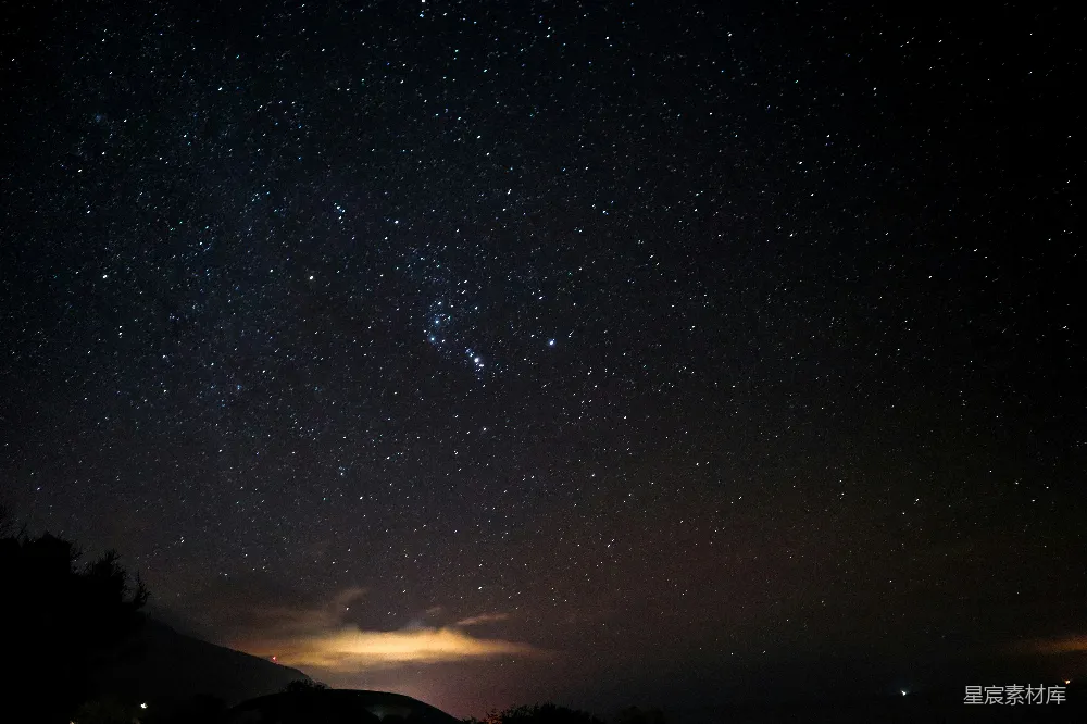 这张图片展示了一个宁静的夜晚，天空中布满了无数闪烁的星星，形成了璀璨的星河。下方的黑暗中隐约可见山脉的轮廓和一些微弱的光点，可能是远处的灯光或城市。整体氛围宁静而神秘，适合用于展示自然美景或夜景主题。