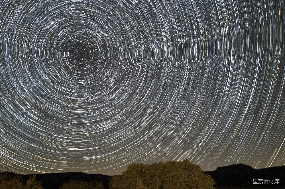 这张图片展示了夜晚星空中的星轨，形成了一种旋转的螺旋图案。下方是树木的剪影，增加了画面的层次感。长曝光技术捕捉到了星星的轨迹，营造出一种梦幻般的氛围。适合用于自然风光、天文摄影或抽象艺术的背景。