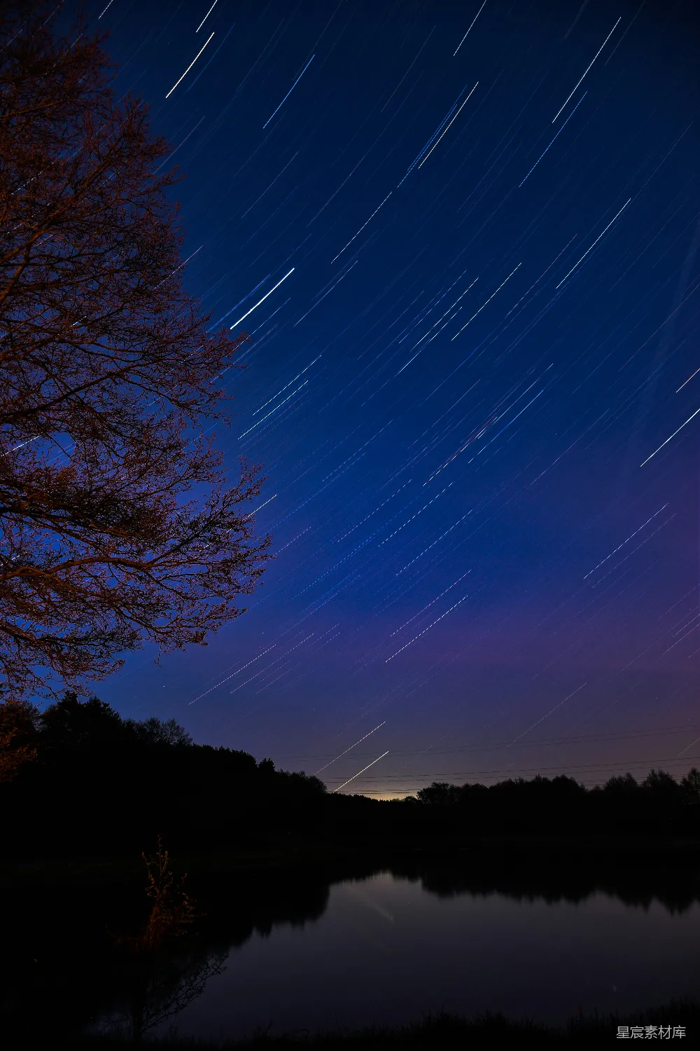 这张图片展示了夜晚湖边的美丽景色，星空中的星星轨迹倒映在平静的湖面上，形成了动与静的完美结合。深邃的蓝色天空与深色的树木和湖面形成了鲜明的对比，营造出一种宁静而神秘的氛围。适合用作自然风光、摄影技巧或宁静夜晚的背景图片。