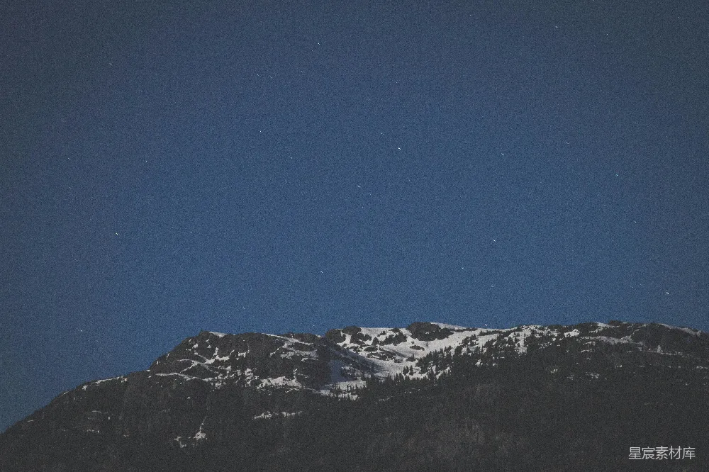 这张图片展示了夜晚时分的雪山景象，天空中布满了星星，山峰上覆盖着白雪，树木在黑暗中若隐若现。整个画面给人一种宁静而神秘的感觉，适合用作自然风光、旅行摄影或冥想背景。