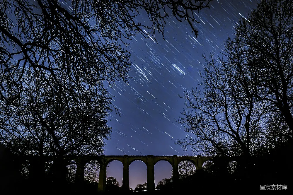 这张图片展示了一座古老的桥梁在夜晚的星空中。桥下是宁静的夜景，星轨的轨迹在夜空中形成了一幅美丽的画面。树木的剪影和桥梁的拱形结构在夜空中形成了鲜明的对比，给人一种宁静而神秘的感觉。适合用于夜景摄影、自然风光、桥梁建筑等主题。