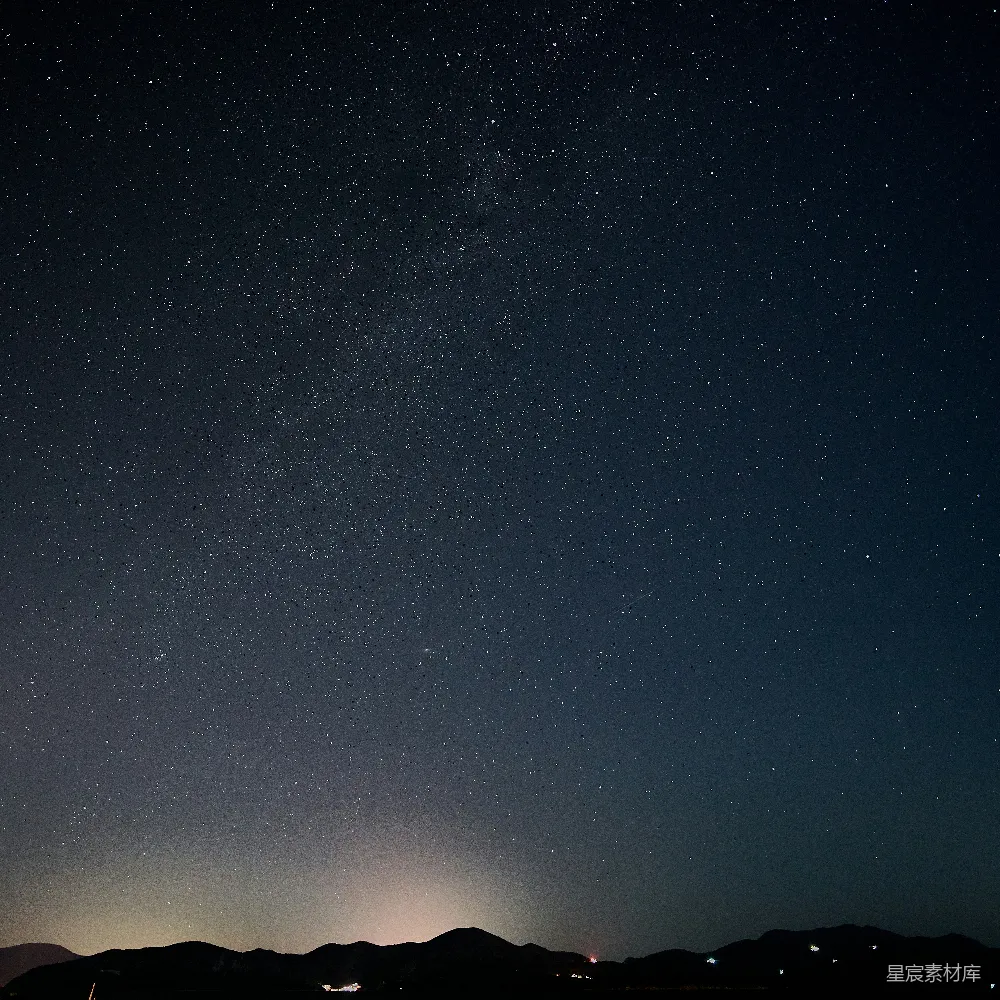 这张图片展示了夜晚的天空，繁星点点，星河横跨天际，下方是轮廓分明的山脉和远处的灯光。这种场景通常适合用于自然风光的摄影，表达宁静与神秘的氛围。