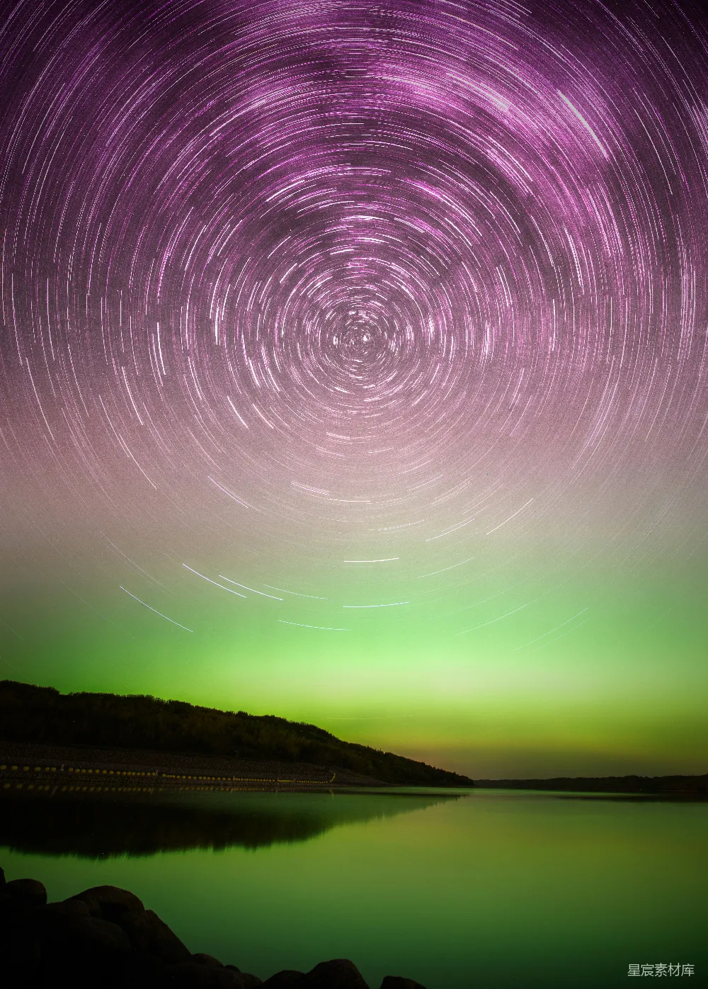 这张图片展示了夜空中繁星点点的星轨和壮观的极光，画面下方是宁静的湖泊和远处的山脉，构成了一幅美轮美奂的自然景观。星轨的旋转和极光的色彩变化，使得整个画面充满了动感与神秘感。适合用作自然风光、摄影艺术和梦幻主题的背景素材。