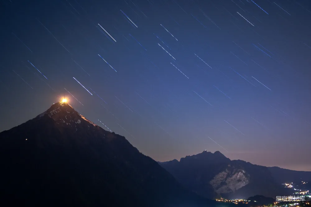 这张图片展示了夜幕下的山峰，星空中的星轨线条清晰，与山峰的轮廓形成鲜明对比。山峰顶部的灯光点缀在黑暗中，营造出一种宁静而神秘的氛围。适合用作自然风光、夜景和宁静主题的背景图片。