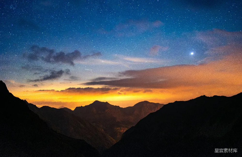 这张图片展示了夜空中繁星点点的壮丽景象，山峦在夜色中形成剪影，呈现出一种宁静而神秘的氛围。天空中橙色的余晖与深邃的蓝色交织，营造出一种梦幻般的视觉效果，适合用作自然风光、旅行或冥想主题的背景。