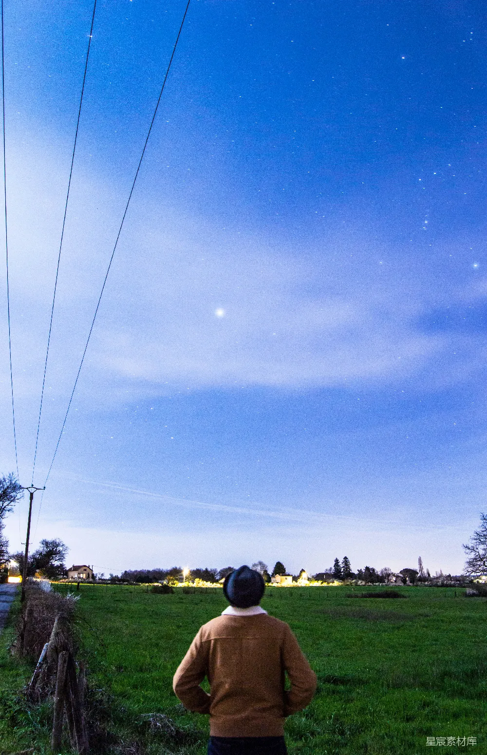 这张照片展示了一个人站在田野上，背对着镜头，仰望星空的场景。天空中布满了星星，地平线远处散布着几座小屋，给人一种宁静而神秘的感觉。照片充满了自然与人文的结合，适合用作旅行、自然风光或人文摄影的素材。