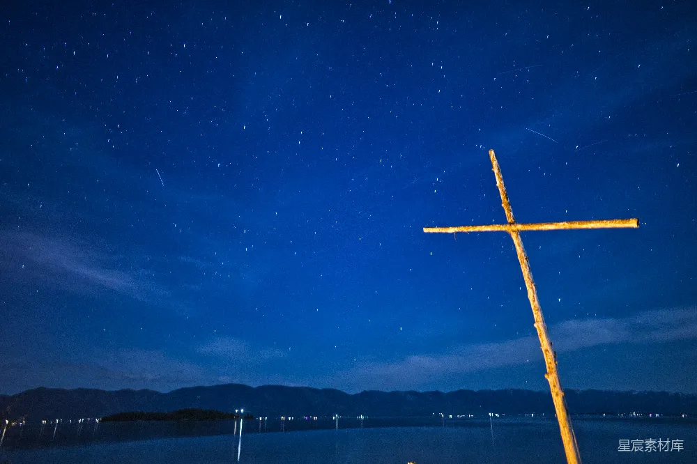 这张图片展示了夜空中布满星星的宁静场景，前景中矗立着一个简单的十字架。背景是远处的山脉和水面上的微弱灯光，营造出一种平和而神秘的氛围。十字架象征着信仰与希望，与美丽的夜空形成对比，让人感受到宁静与深邃。