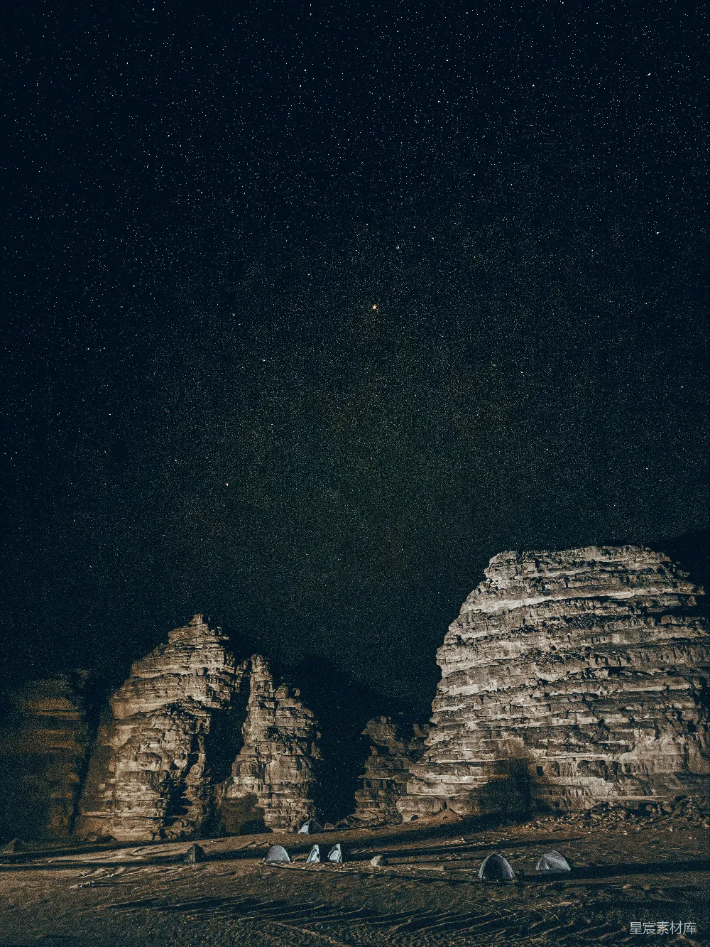 这张图片展示了一群人在星空下露营的场景。夜空中布满了星星，岩石在黑暗中显得格外神秘。露营者在岩石之间搭建了帐篷，享受着宁静的夜晚。适合用于自然风光、户外活动、星空摄影等场景。