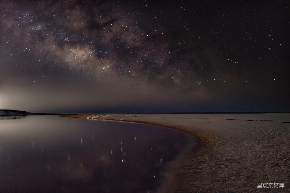 这张图片展示了一片宁静的海滩，银河横跨夜空，星星点缀其间。海面反射出夜空的景象，形成了一幅美丽的倒影画面，给人一种宁静而神秘的感觉。适合用于展示自然美景、夜景摄影或旅行景点宣传。