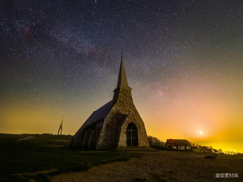 这张图片展示了在夜空下宁静的教堂场景。星空璀璨，教堂在微弱的灯光下显得格外突出，背景中可以看到远处的建筑和树木。整体氛围宁静而神秘，适合用作自然风光、宁静生活或宗教主题的背景素材。
