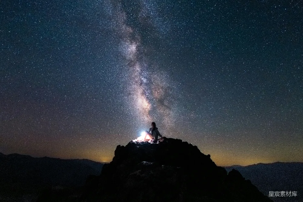 这张图片展示了一位坐在山巅上的人，仰望着璀璨的星空。银河横跨夜空，星星点缀其间，营造出一种宁静而神秘的氛围。背景是连绵的山脉，远处的天空渐变至深蓝，给人一种远离尘嚣的感觉。这幅图像适合用作自然风光、旅行和摄影爱好者的素材。