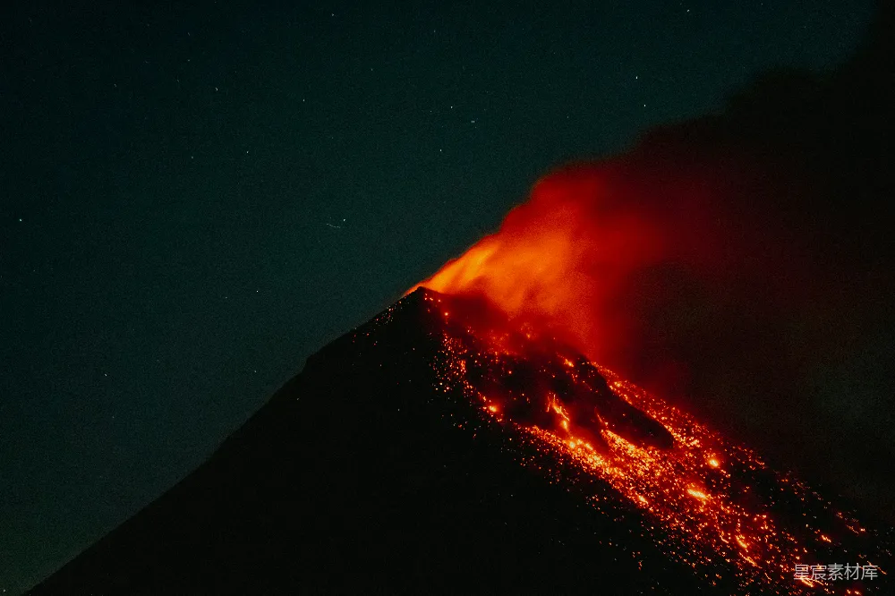 这幅图片展示了夜晚火山爆发的壮观景象。火山口喷发出炽热的熔岩和烟雾，映衬在夜空中，形成鲜明的橙红色光芒。场景中火山灰和熔岩流沿着山坡向下蔓延，展示了自然力量的震撼力量。适合用作自然、地质或灾害主题的背景素材。