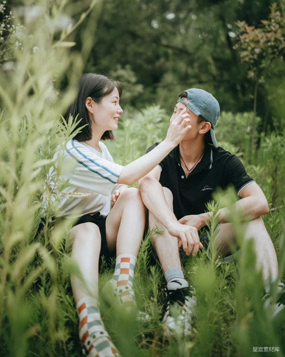 这张图片展示了一对情侣在茂密的草地上温馨互动的场景。女性轻柔地抚摸着男性的脸颊，两人面带微笑，显得十分甜蜜。背景是郁郁葱葱的绿色植物，增添了画面的自然气息。这张照片适合用于展示爱情故事、自然风光或是情侣写真。