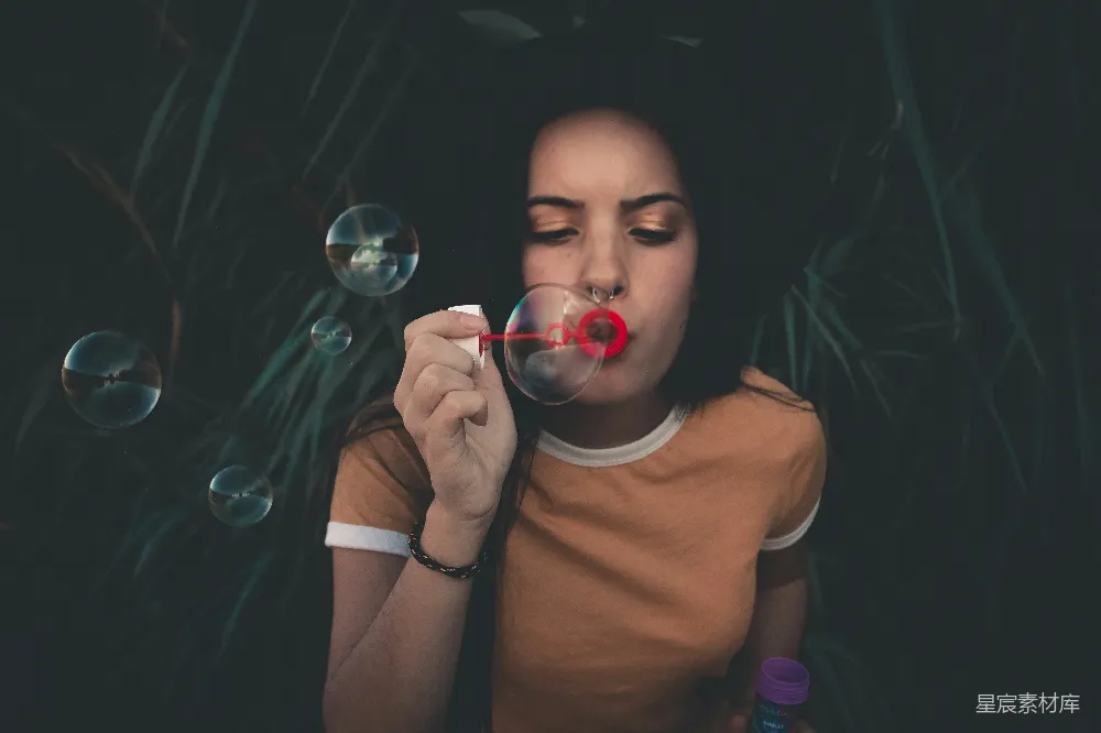 图片中，一位女孩正在户外吹泡泡，背景是茂密的植物。她专注地吹着泡泡，手中握着泡泡棒，泡泡在空中飘散，营造出一种宁静而美好的氛围。照片的色调柔和，自然的光线和植物的影子为画面增添了深度和层次感。适合用作自然、休闲、童趣相关的背景图。