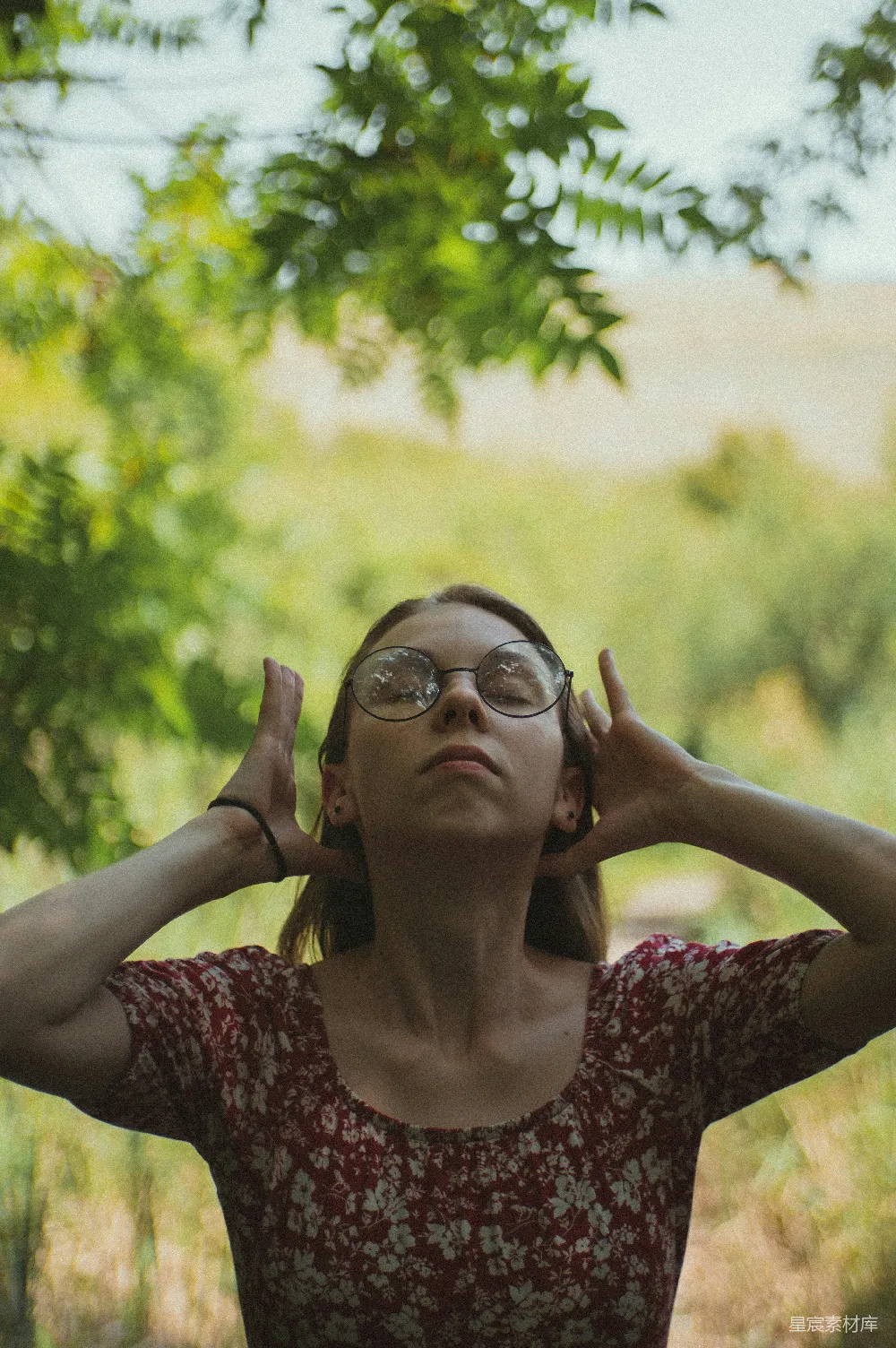 图片中，一位女士在夏日的树荫下，穿着红色的花卉连衣裙，双手轻轻托住头部，似乎在享受片刻的宁静与放松。背景是模糊的绿树和柔和的阳光，营造出一种自然、宁静的氛围。这幅画面适合用于表现夏日户外休闲、自然放松的主题。