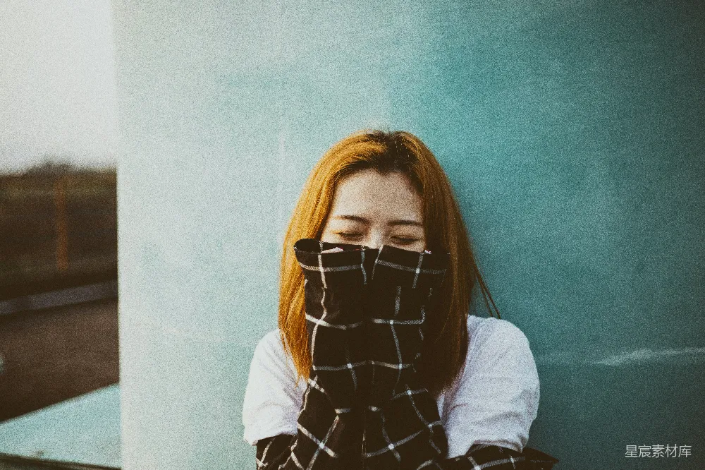 一位年轻女子在城市的街头，手持一块格子布遮住了下半张脸，她站在一扇蓝色的窗户前，背景略显朦胧，给人一种朦胧和宁静的感觉。这种街头风格的肖像捕捉到了现代都市生活中年轻人的瞬间，传达出一种独特的时尚和情感氛围。