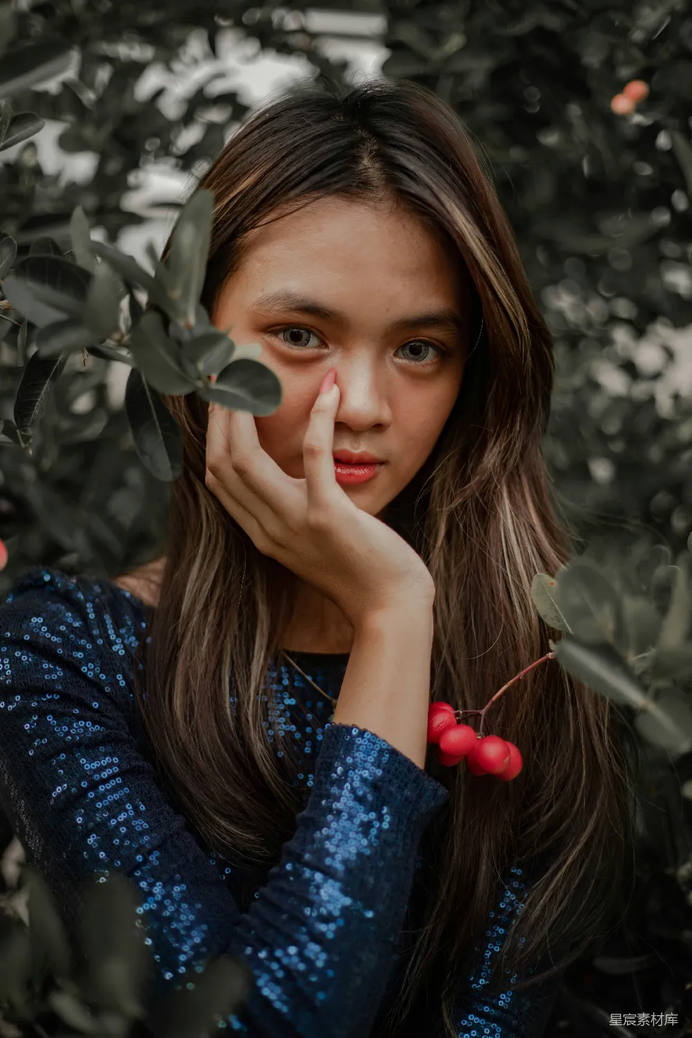这是一张拍摄于自然环境中的照片，一位女孩在树叶和红色浆果的衬托下，用手指轻触脸颊，营造出一种宁静而自然的氛围。她穿着闪亮的深蓝色上衣，与周围的绿色植物形成鲜明对比，给人一种清新脱俗的感觉。这张照片适合用作自然风光、人物摄影或宁静生活主题的背