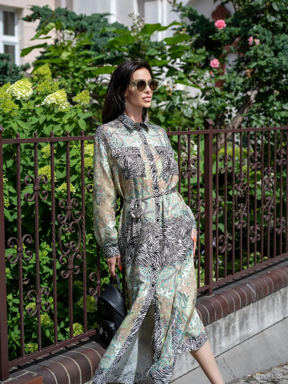 图片中一位女性身穿充满图案设计的长袖衬衫，搭配墨镜，漫步在绿意盎然的花园中。背景是铁艺栏杆和盛开的花朵，营造出一种悠闲自在的氛围。整体风格时尚而自然，适合用于展示休闲风格的服装或户外活动场景。