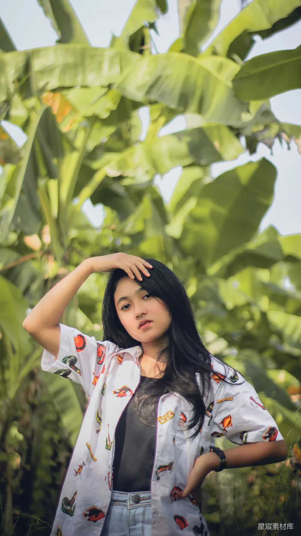 这张图片展示了一位年轻女孩在蕉叶下摆姿势的场景，背景是茂密的绿色蕉叶，阳光透过叶子洒在女孩的身上，营造出一种宁静而自然的氛围。女孩穿着印有图案的衬衫，显得既时尚又自然。整个画面充满了青春的气息和自然的美感，适合用作自然风光、青春写真或户外摄