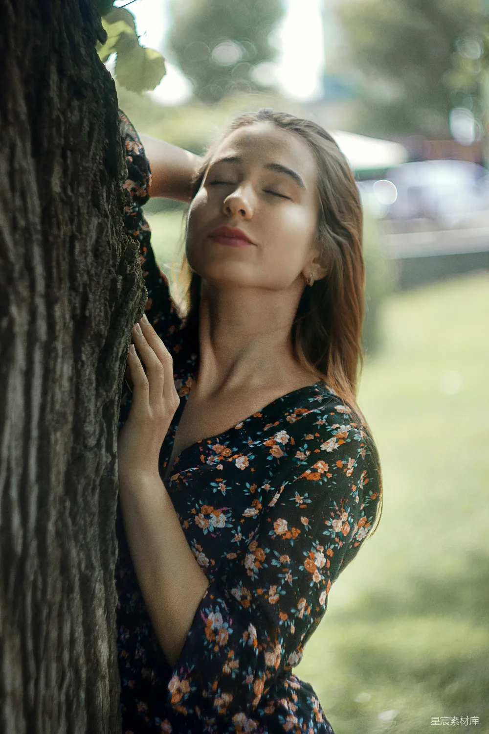 图片展示了一位穿着花卉连衣裙的少女，她靠在一棵树上，闭着眼睛，似乎在享受大自然的宁静。背景是模糊的绿色植物和自然光，营造出一种宁静而平和的氛围。适合用于表达自然、宁静、女性肖像等主题。