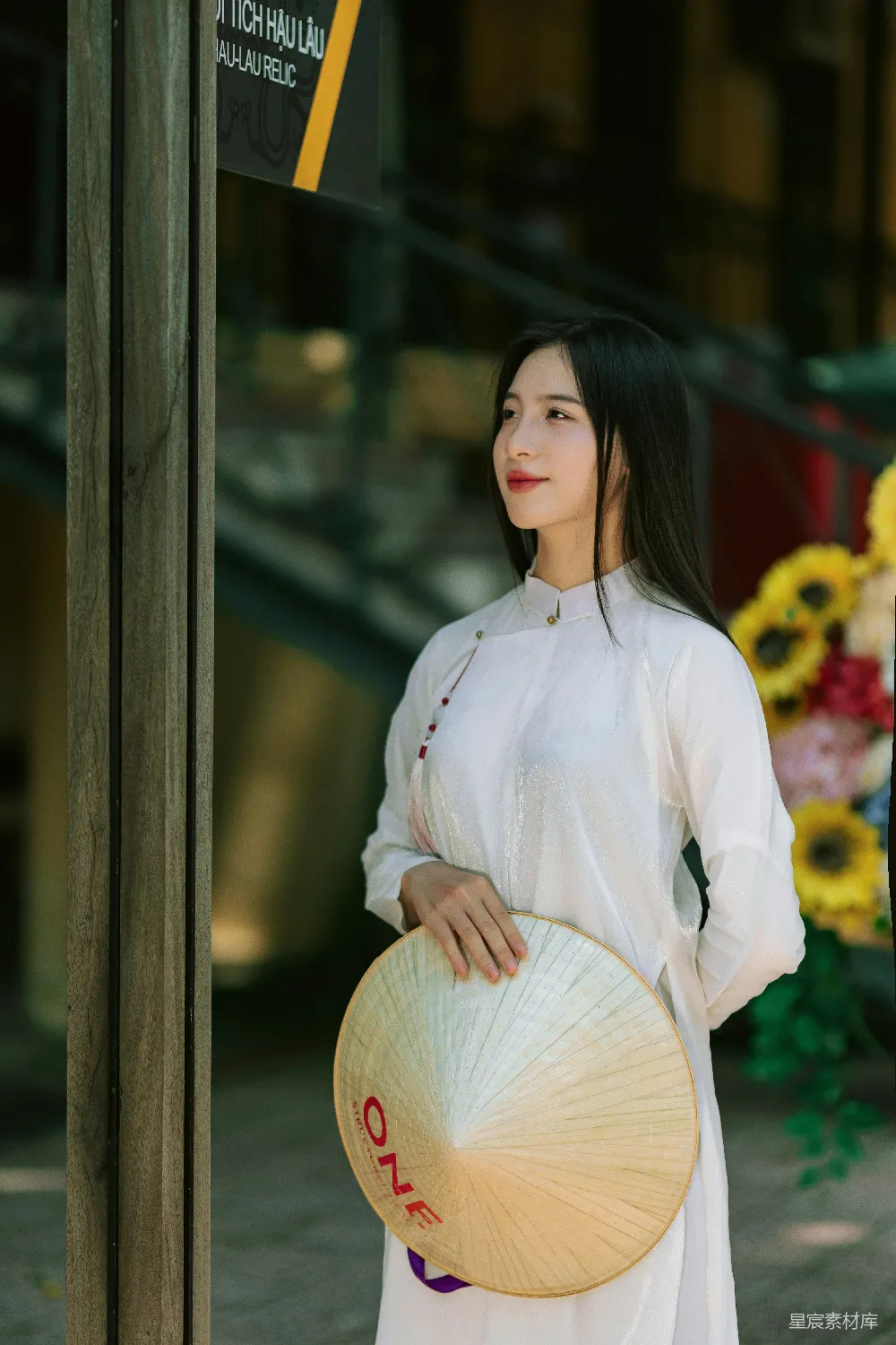一位身着传统越南旗袍的女性，手握一顶精致的竹编斗笠，静立于一扇古色古香的门前，背景中隐约可见的向日葵与木制结构增添了自然与质朴的氛围。这幅画面不仅展现了越南独特的文化韵味，还捕捉到了一种宁静而优雅的生活场景。