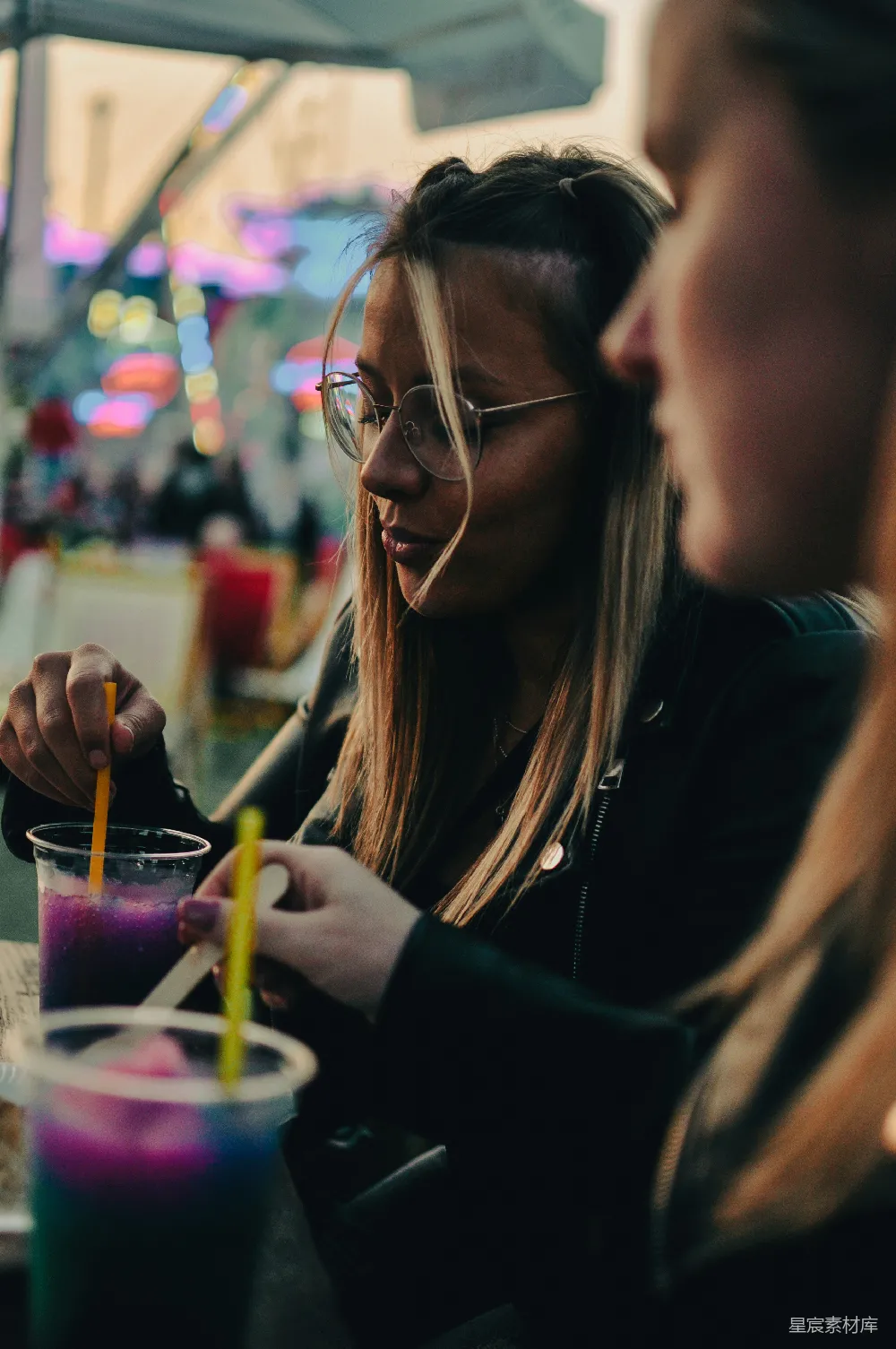 图片中，一位长发女性正在夜市中与朋友分享一杯色彩斑斓的饮料，背景的霓虹灯增添了夜晚的热闹氛围。这是一段放松和享受的时光，适合用在描述朋友聚会或夜生活场景中。