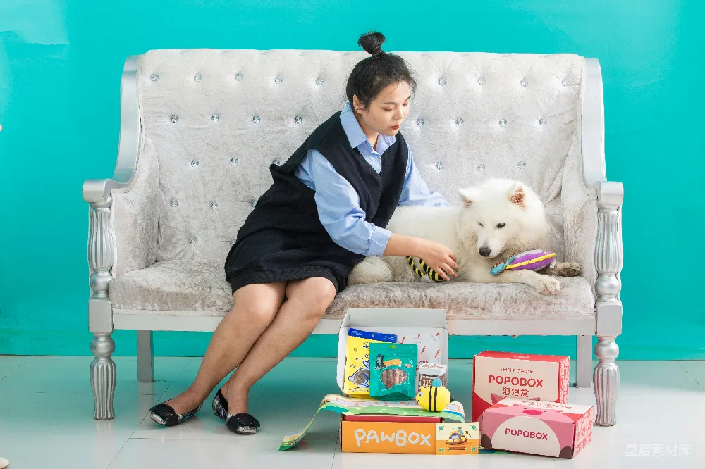图片中，一位年轻女性与一只可爱的萨摩耶犬坐在装饰精美的沙发上，周围摆放着几个宠物零食和玩具的盒子。背景是明亮的蓝色，整体色调和谐温馨，适合表现宠物与主人的日常生活场景，传达出家庭和宠物之间的亲密关系。
