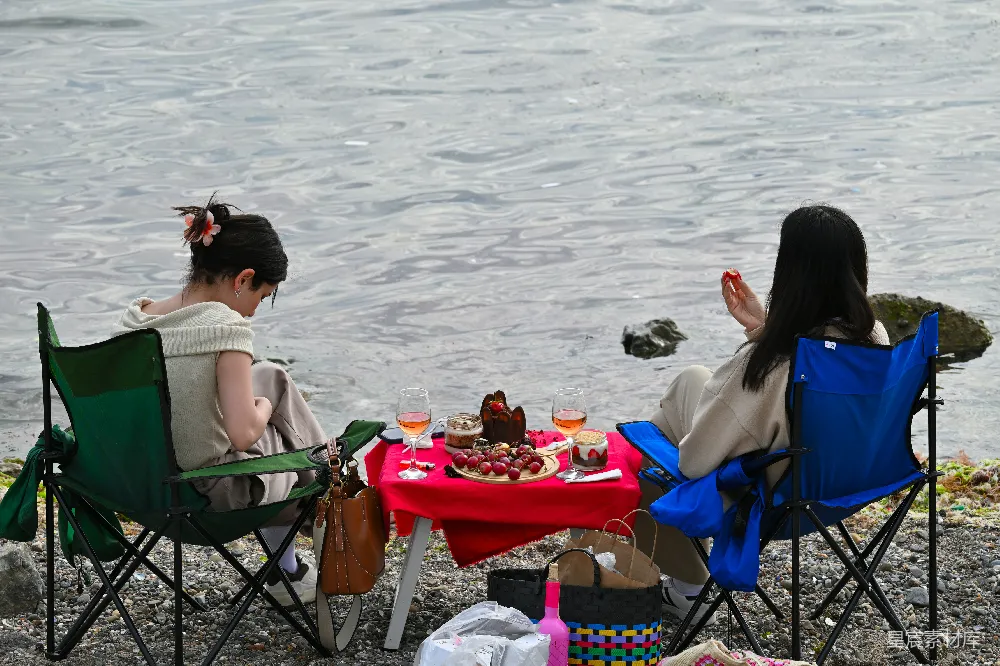 这张图片展示了两位女性在湖畔进行野餐的场景。她们坐在折叠椅上，面前是一张铺着红色桌布的小桌子，上面摆放着各种美食和饮料，如水果、蛋糕和葡萄酒。背景是波光粼粼的湖水，周围是岩石和沙滩，营造出一种宁静而浪漫的氛围。这张图片适合用于户外休闲、野餐